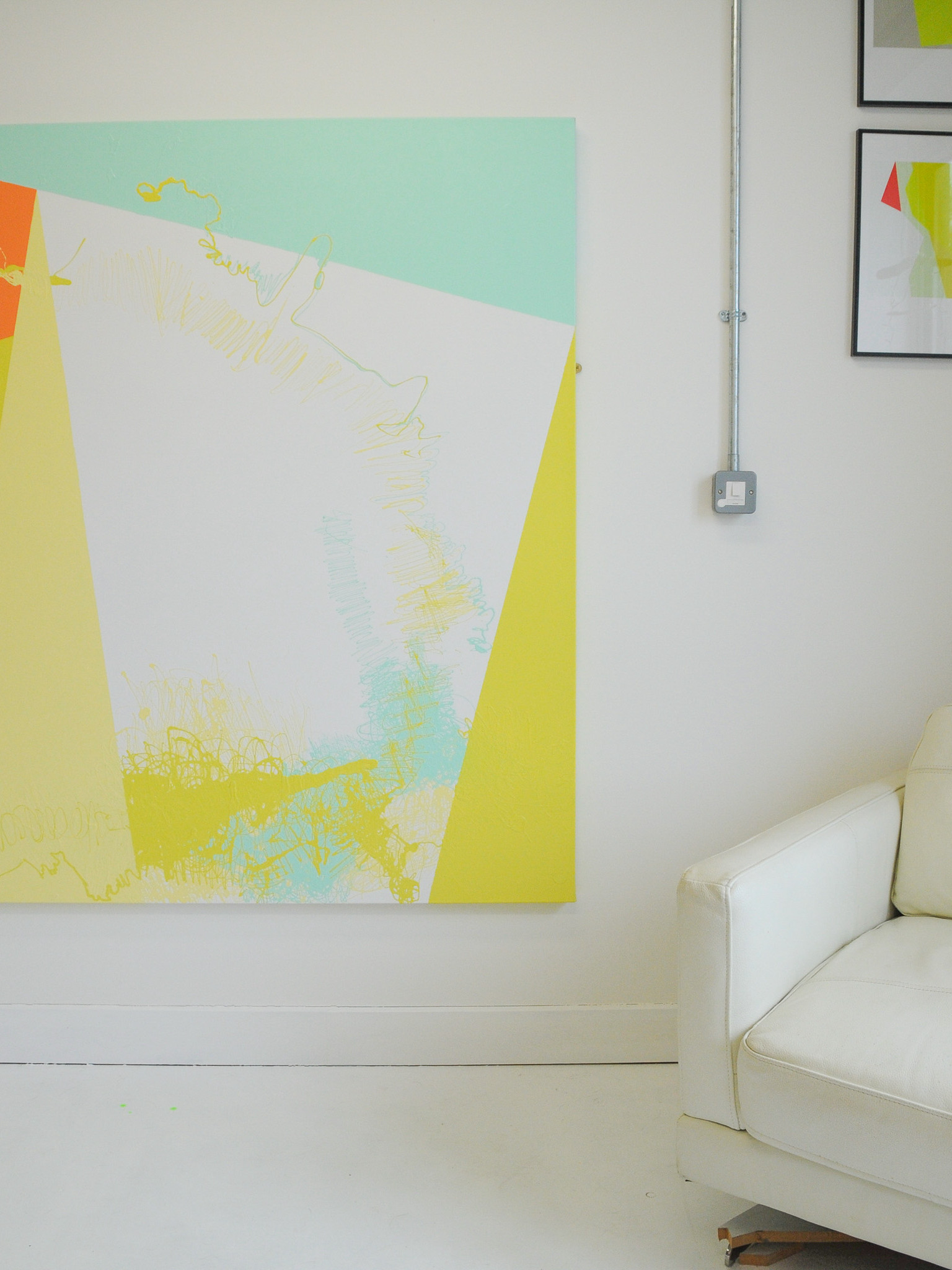 large abstract canvas in geometrics, pastel yellow white and spearmint hues sits in a white room with a white chair and framed prints to the side