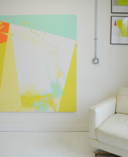 large abstract canvas in geometrics, pastel yellow white and spearmint hues sits in a white room with a white chair and framed prints to the side